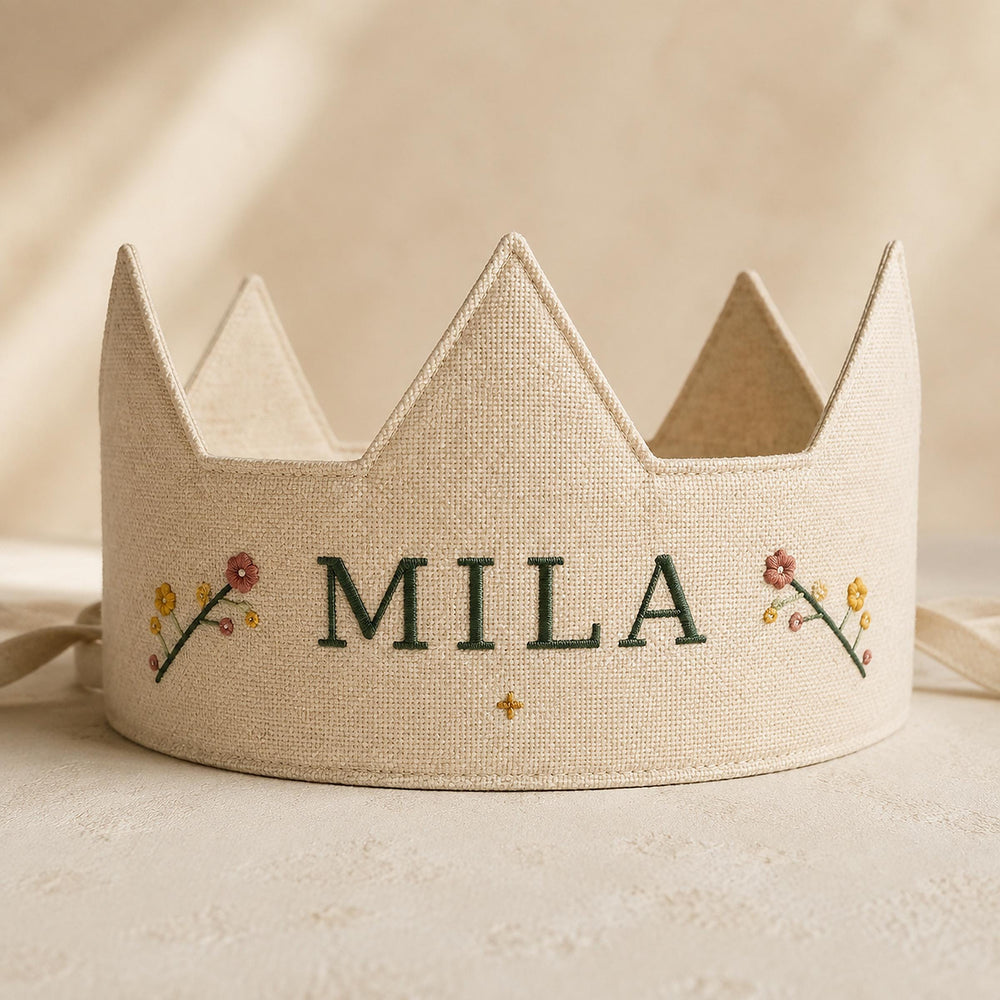 A beige crown with the word "MILA" embroidered on it, along with small floral designs on either side.